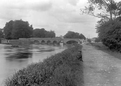 Black and white photo of horse bridge with six arches taken from west bank of river. Boy fishing from the towpath.