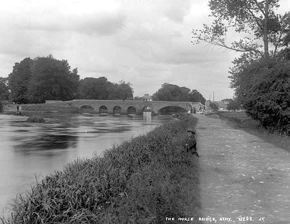 Black and white photo of horse bridge with six arches taken from west bank of river. Boy fishing from the towpath.