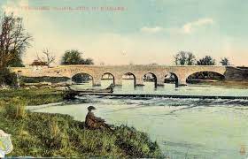 Colourised photo of horse bridge with six arches and weir. Boy fishing in front of weir.